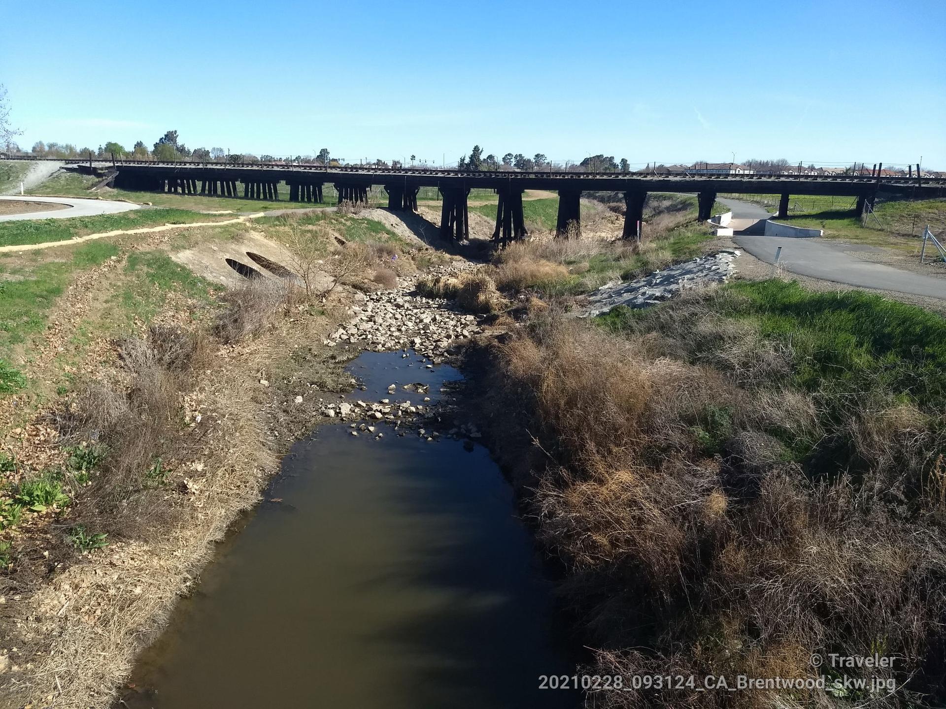 A stream flows under a long wooden bridge on a bright day.