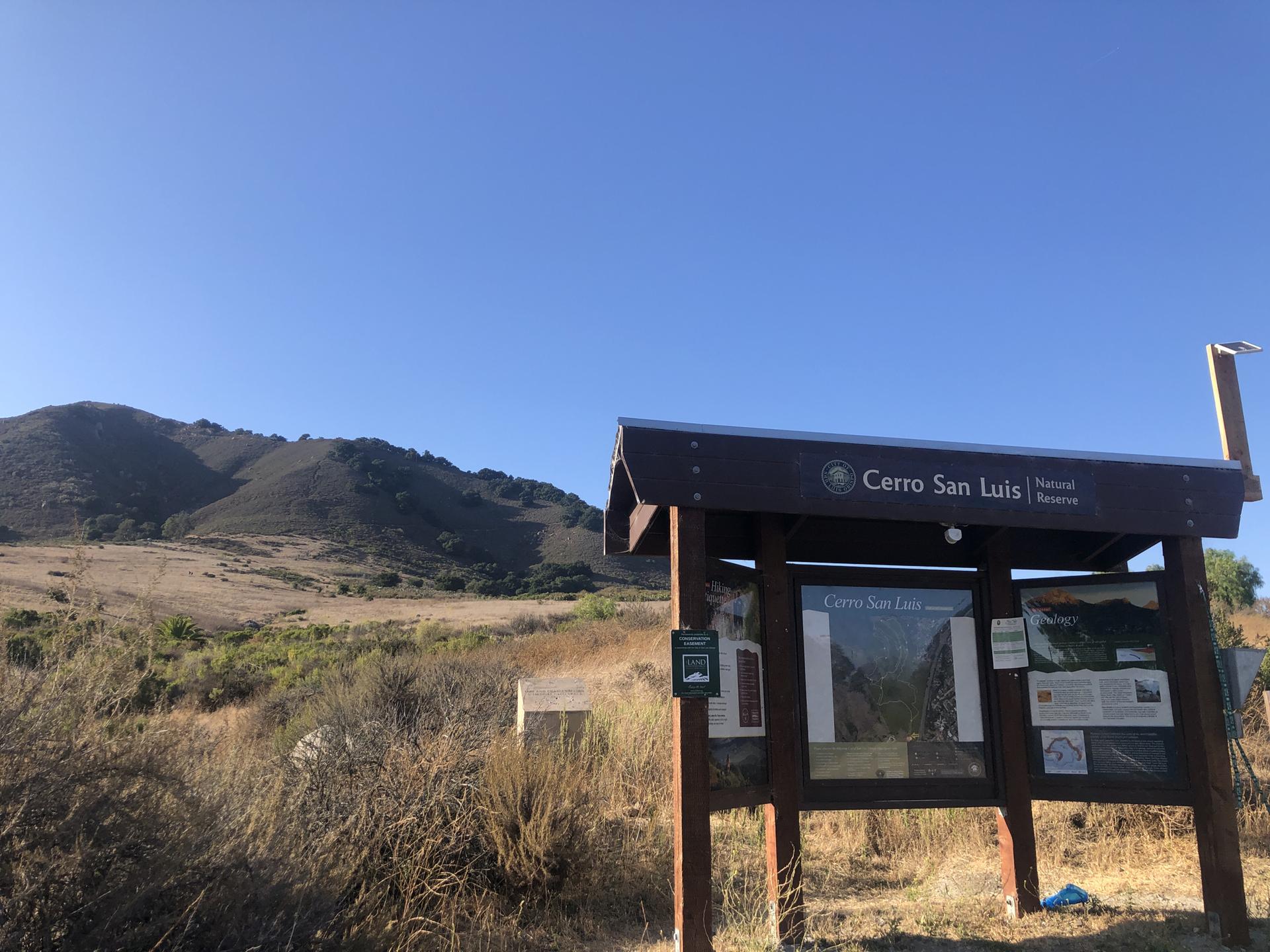 A wooden information sign stands in dry grass before sunny hills.