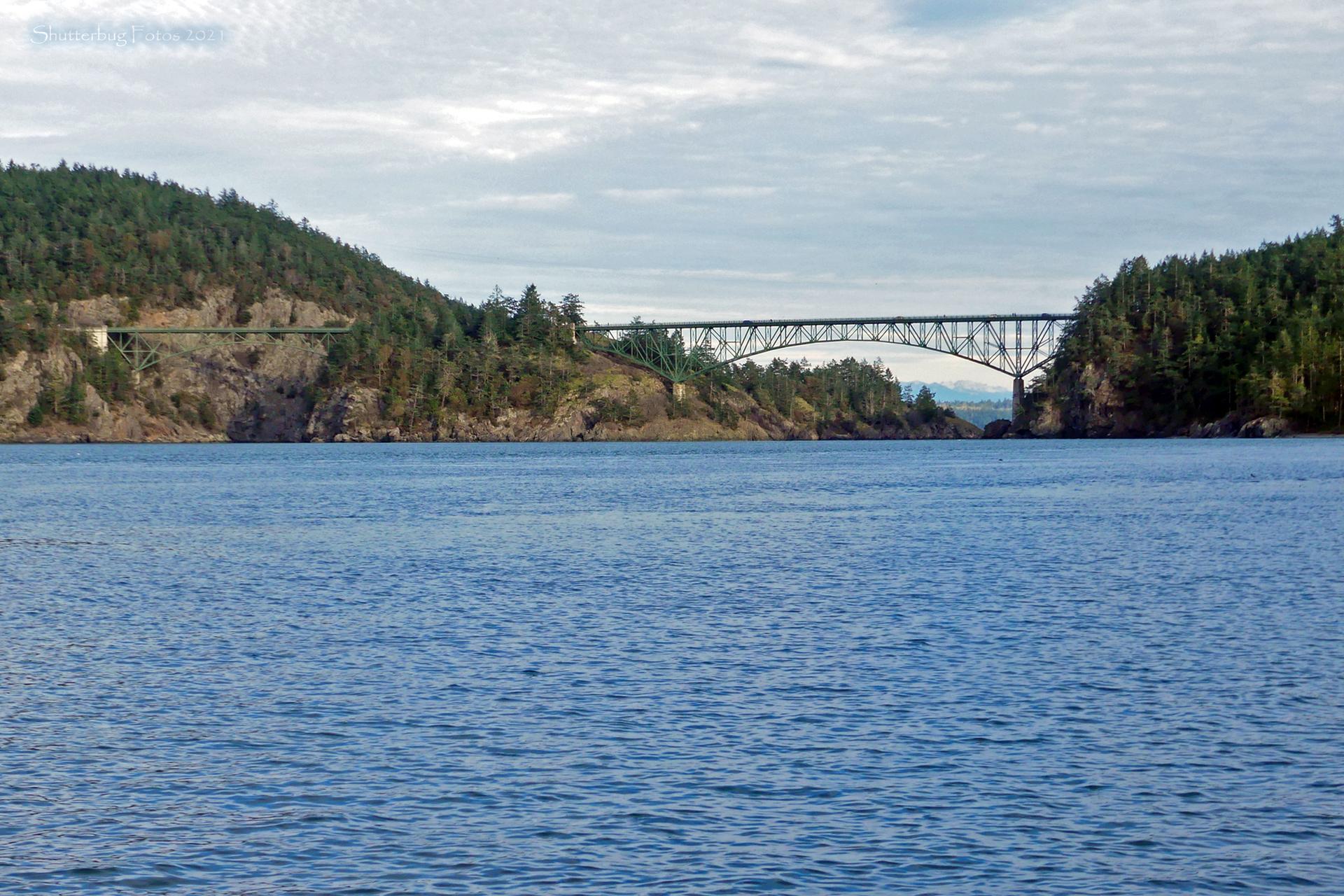 A large steel bridge spans blue water between two forested hills.