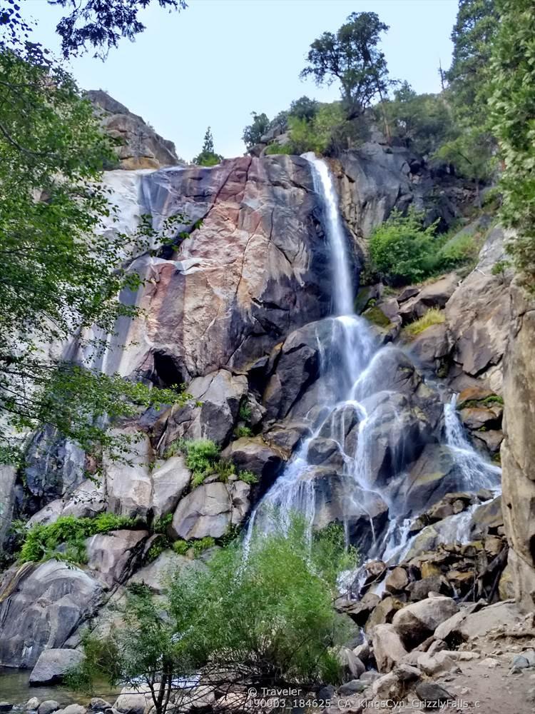 Tall waterfall cascades down a rocky cliff, framed by green trees.