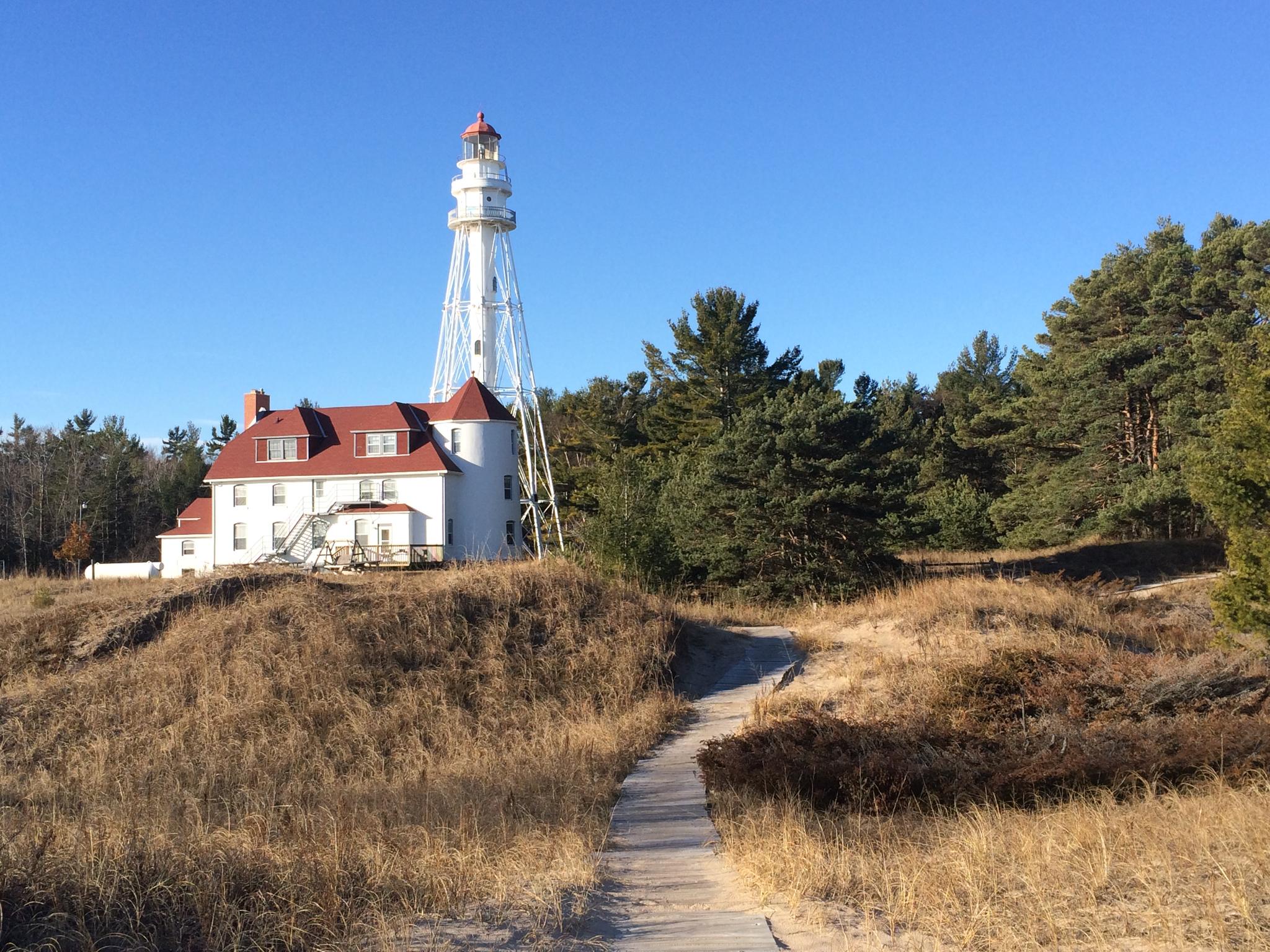 White lighthouse and red-roofed building on a grassy hill with a boardwalk path.