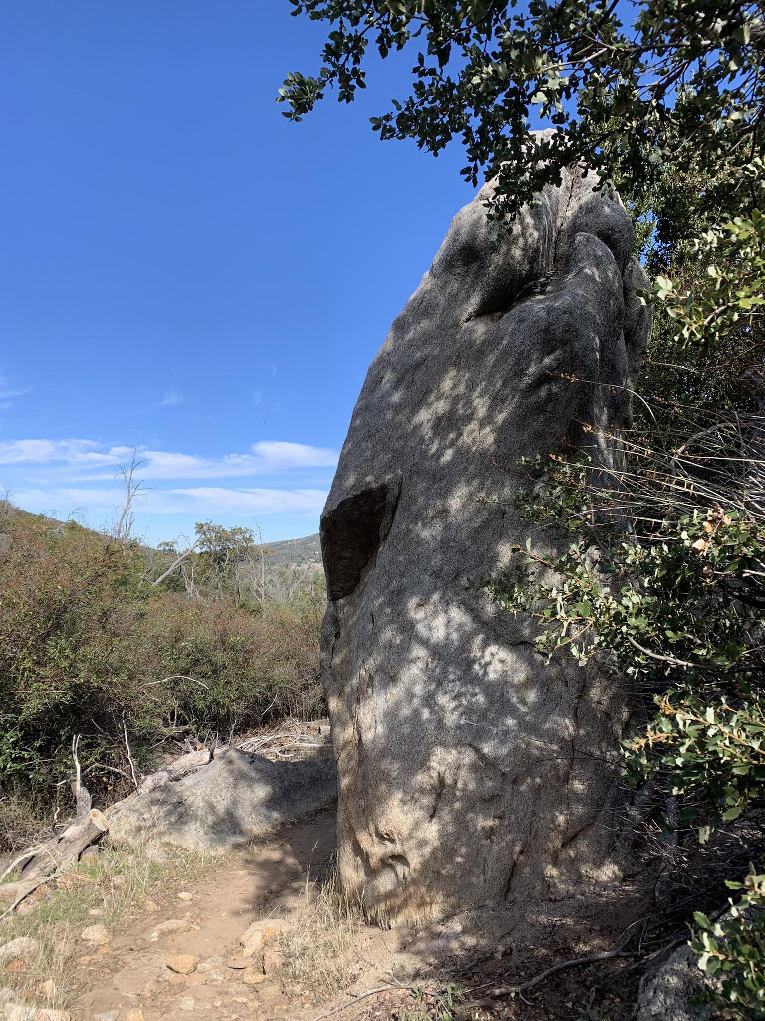 Tall, weathered rock formation beside a dirt path, dappled in sunlight.