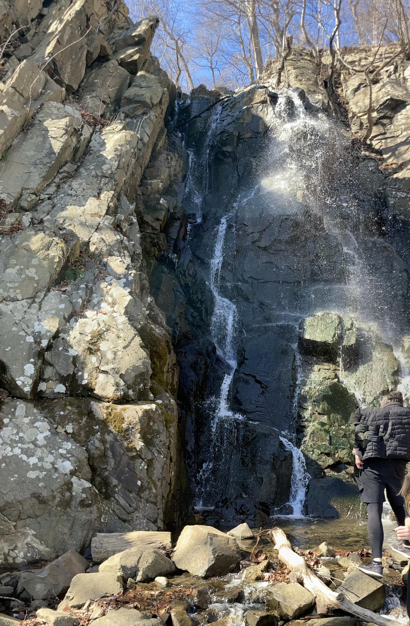 Person standing near a tall waterfall cascading down a sunny, rocky cliff.