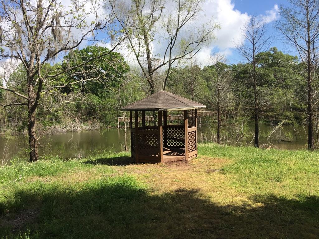 A wooden gazebo sits on a grassy bank beside a calm body of water.