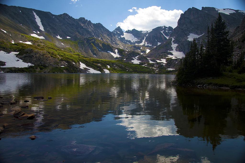 Sunny alpine lake reflecting clouds, surrounded by snow-dusted mountains and green slopes.