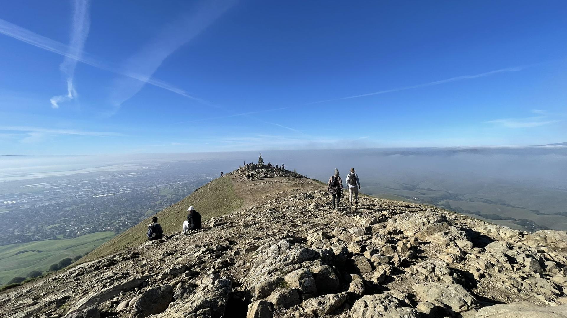 Hikers on a rocky mountain peak overlooking a city and distant fog.