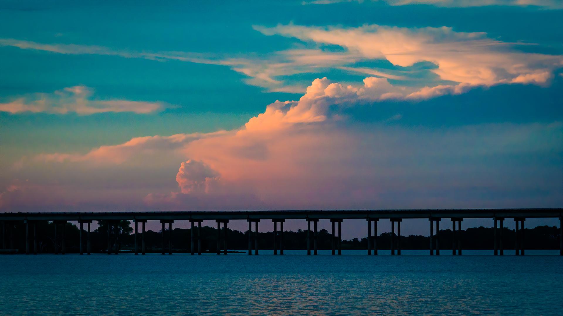 Silhouetted bridge over water with colorful clouds at sunset.