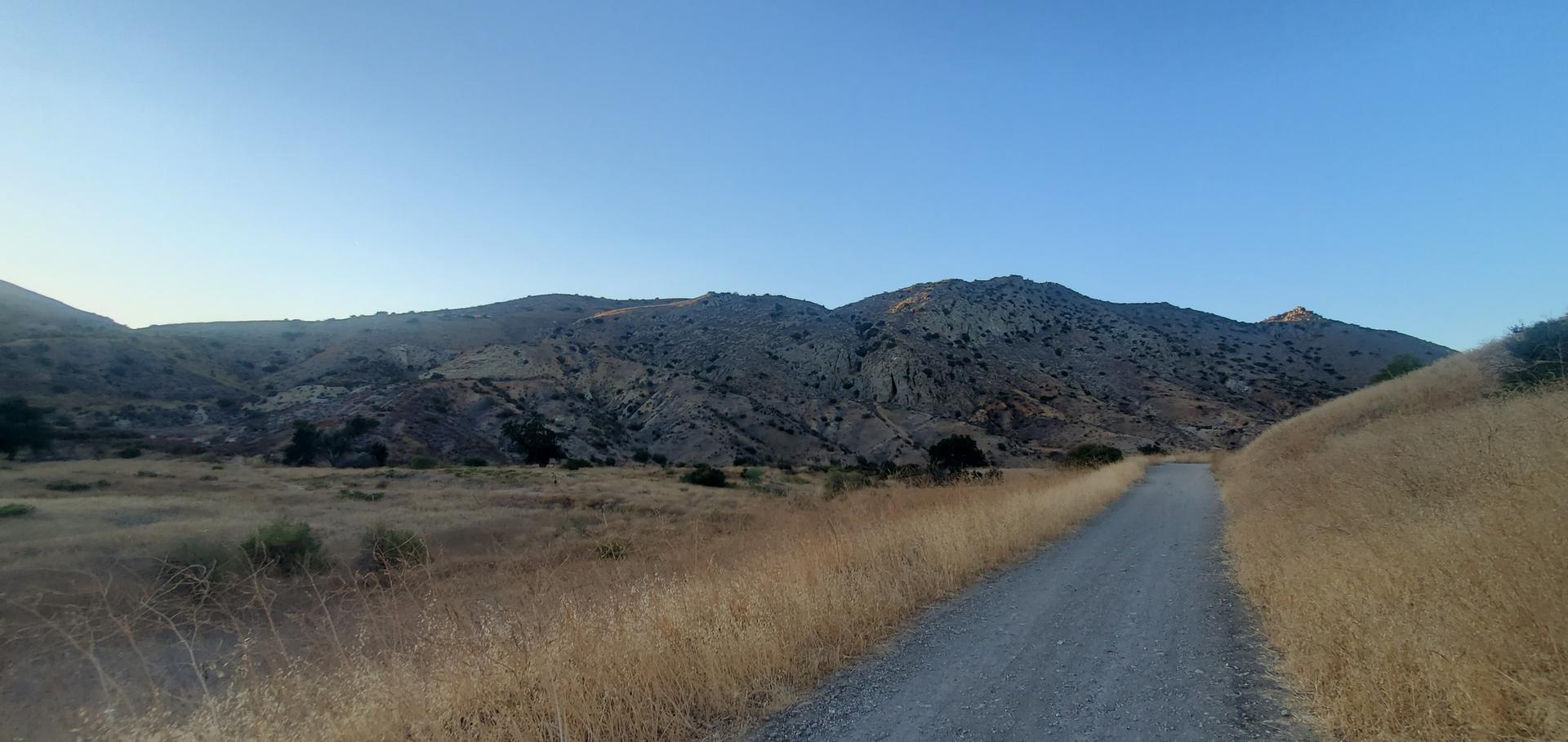 Dirt road through dry grass fields with rugged mountains under a clear sky.