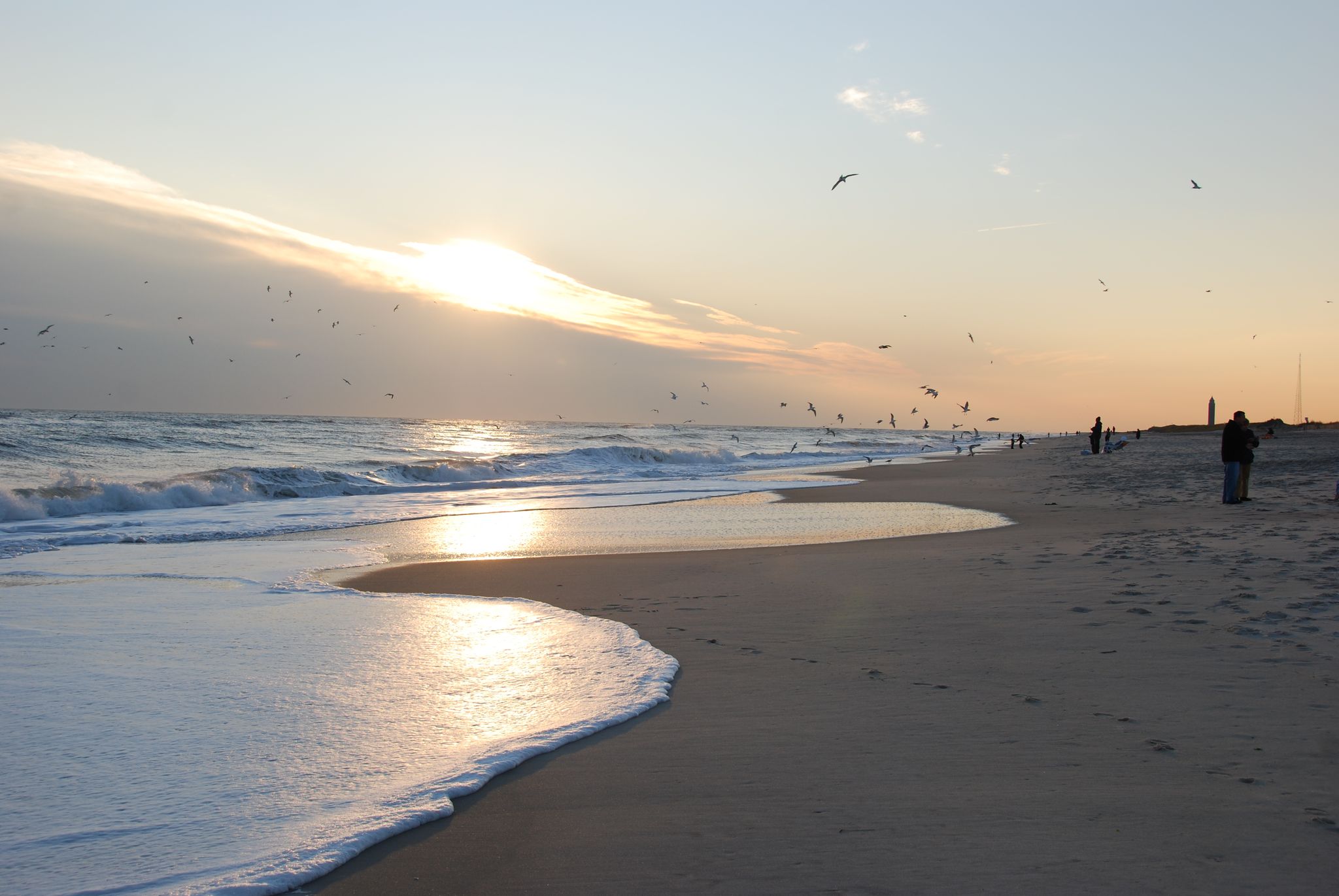 Golden sunset over a sandy beach with waves, many birds flying, and people. - Ocean Parkway Coastal Greenway