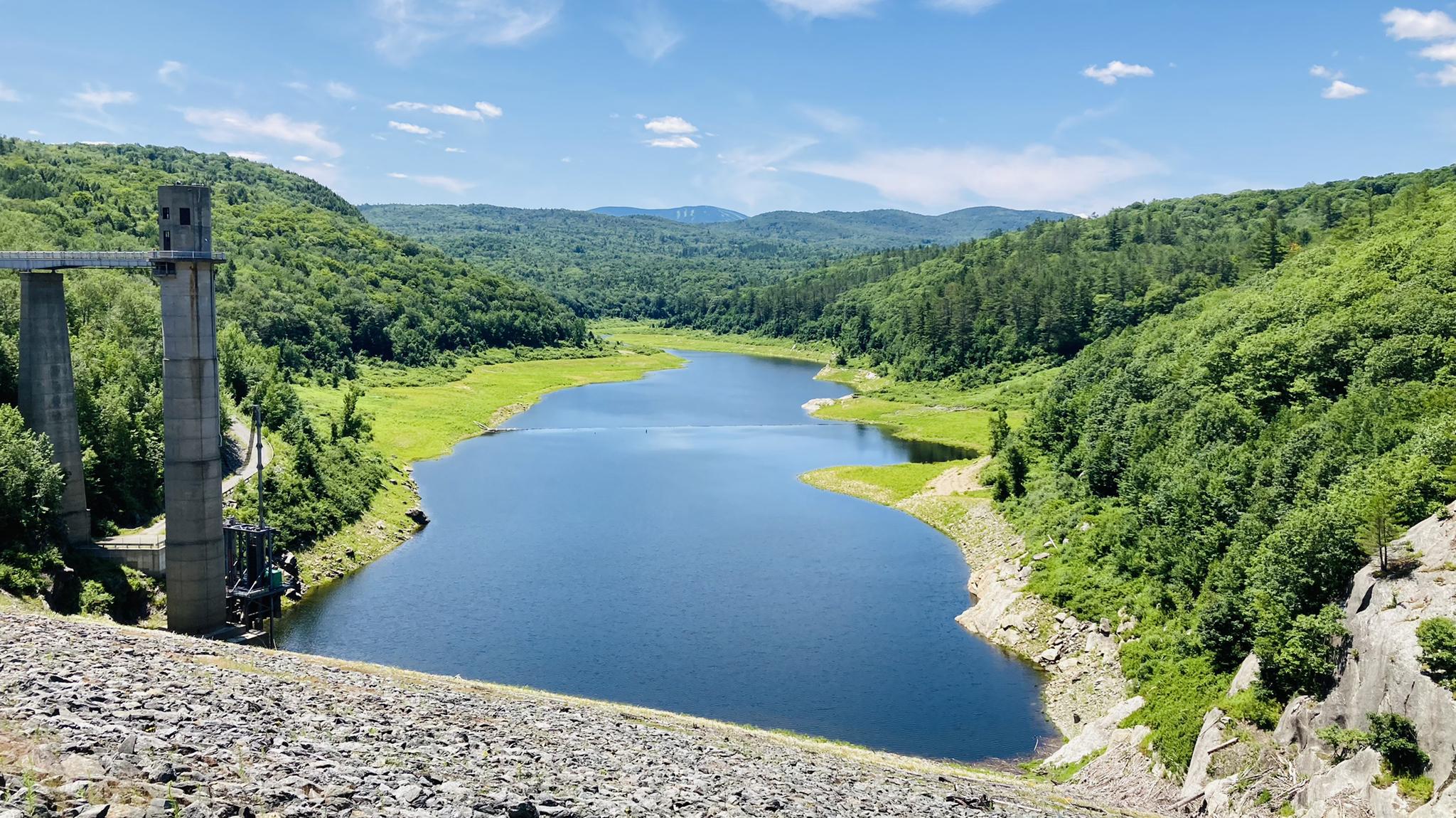 A reservoir with a concrete structure, surrounded by green forested hills under a blue sky.