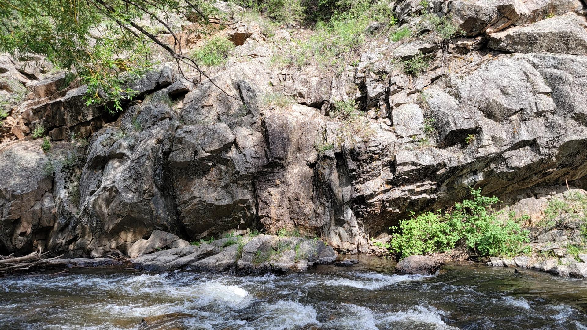 A rushing river flows past a tall, rugged rock cliff in bright sunlight.