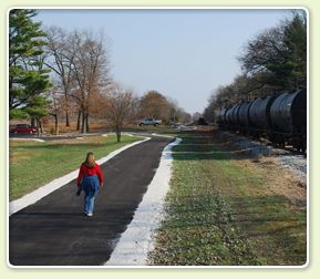 Person walks on a paved path beside parked train cars on a clear day. - Kingsbury, Knox & North Judson loop from Wanatah
