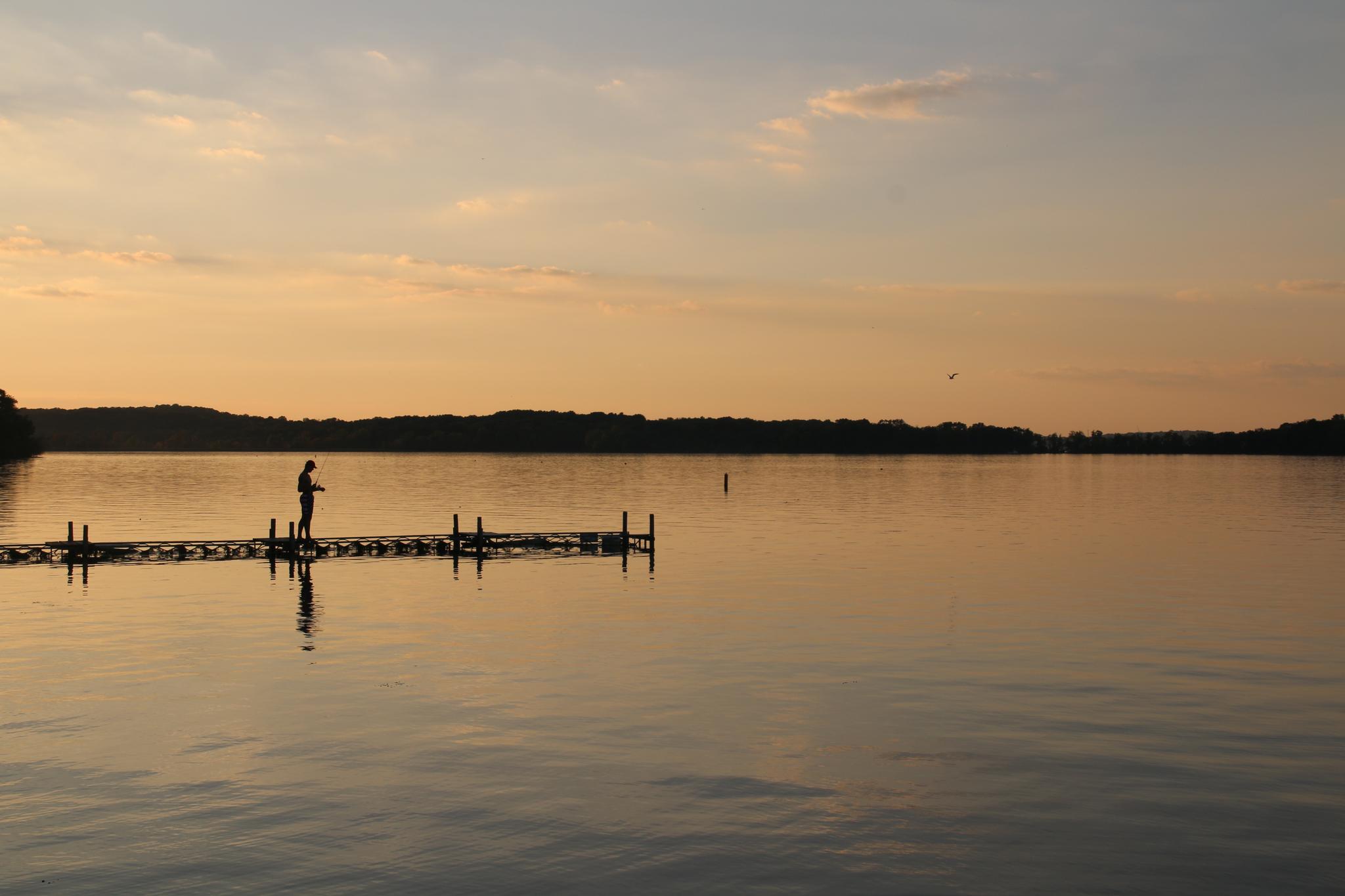 Silhouetted person on a dock over a calm lake at golden hour sunset.