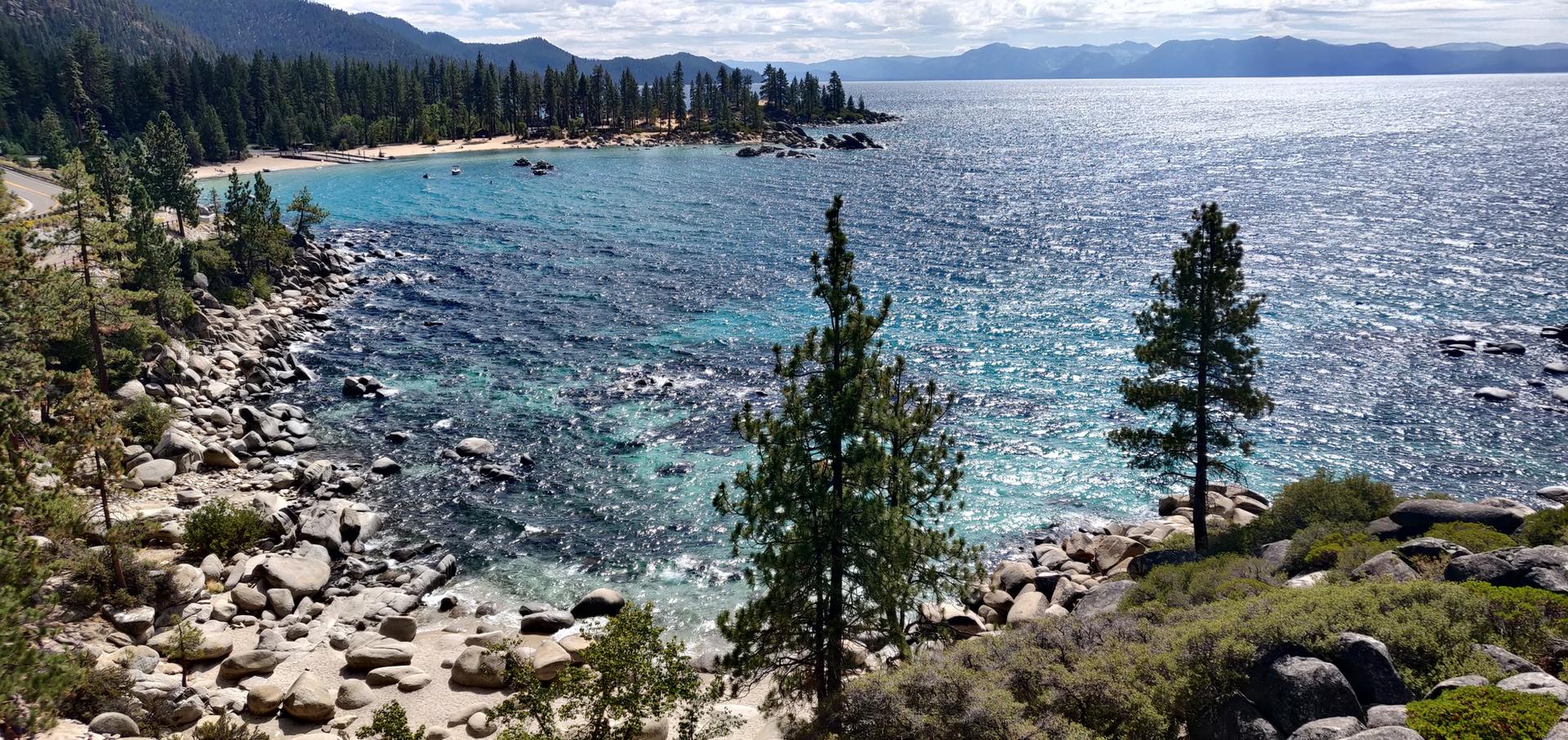 Panoramic view of a clear blue lake, rocky shore, and forested mountains.