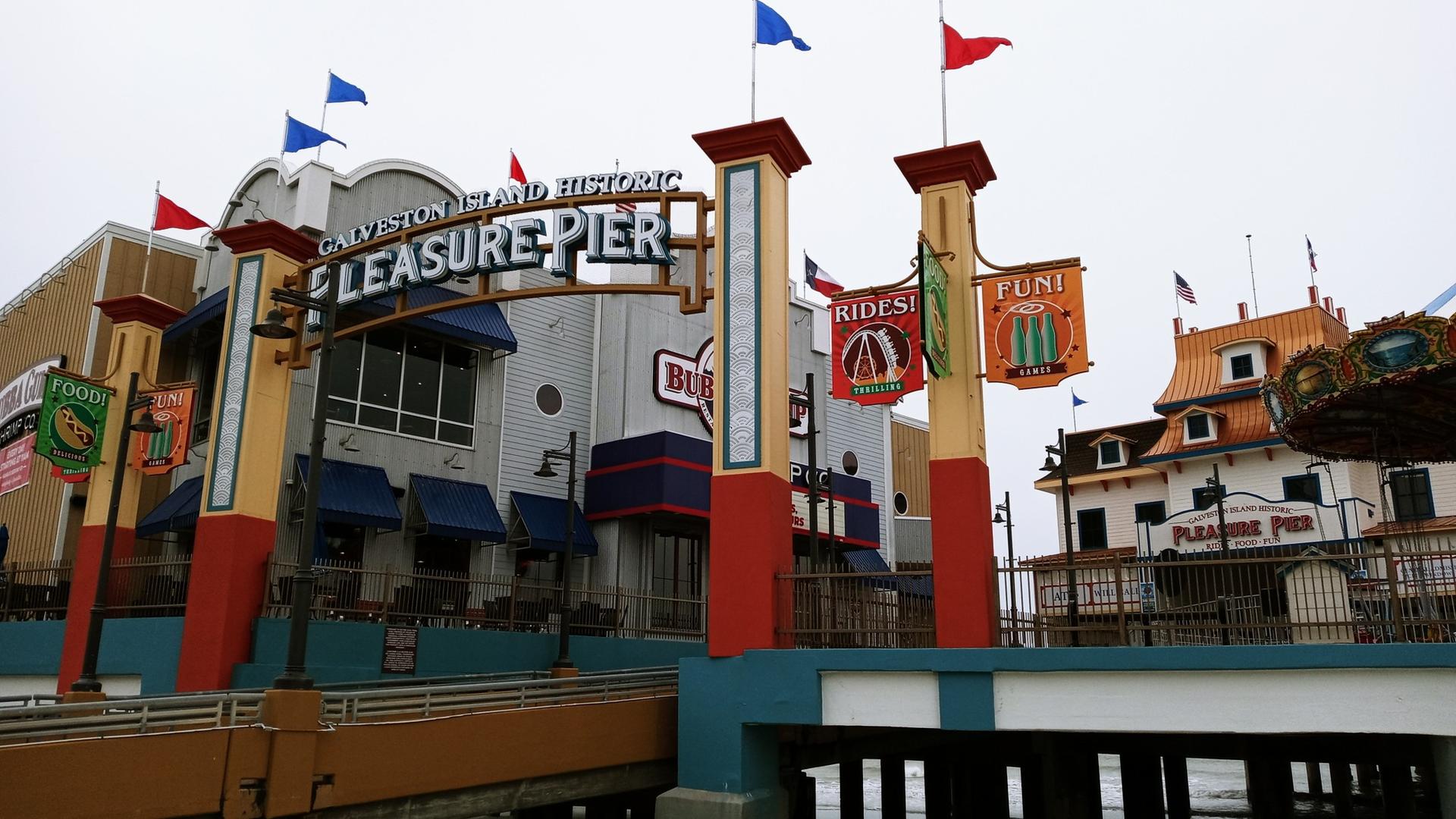 Colorful amusement pier entrance with signs, flags, and a carousel under an overcast sky.