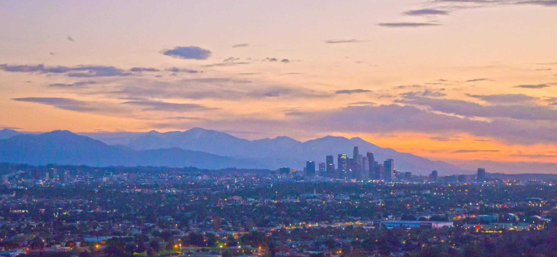 A city skyline and distant mountains at sunset with orange and purple clouds.
