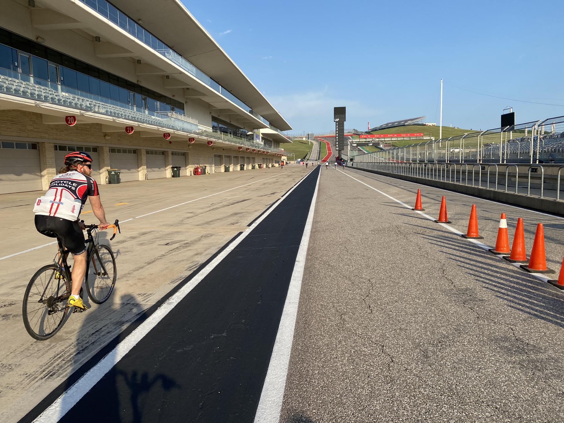 A person cycles on a wide paved track with buildings and orange cones.