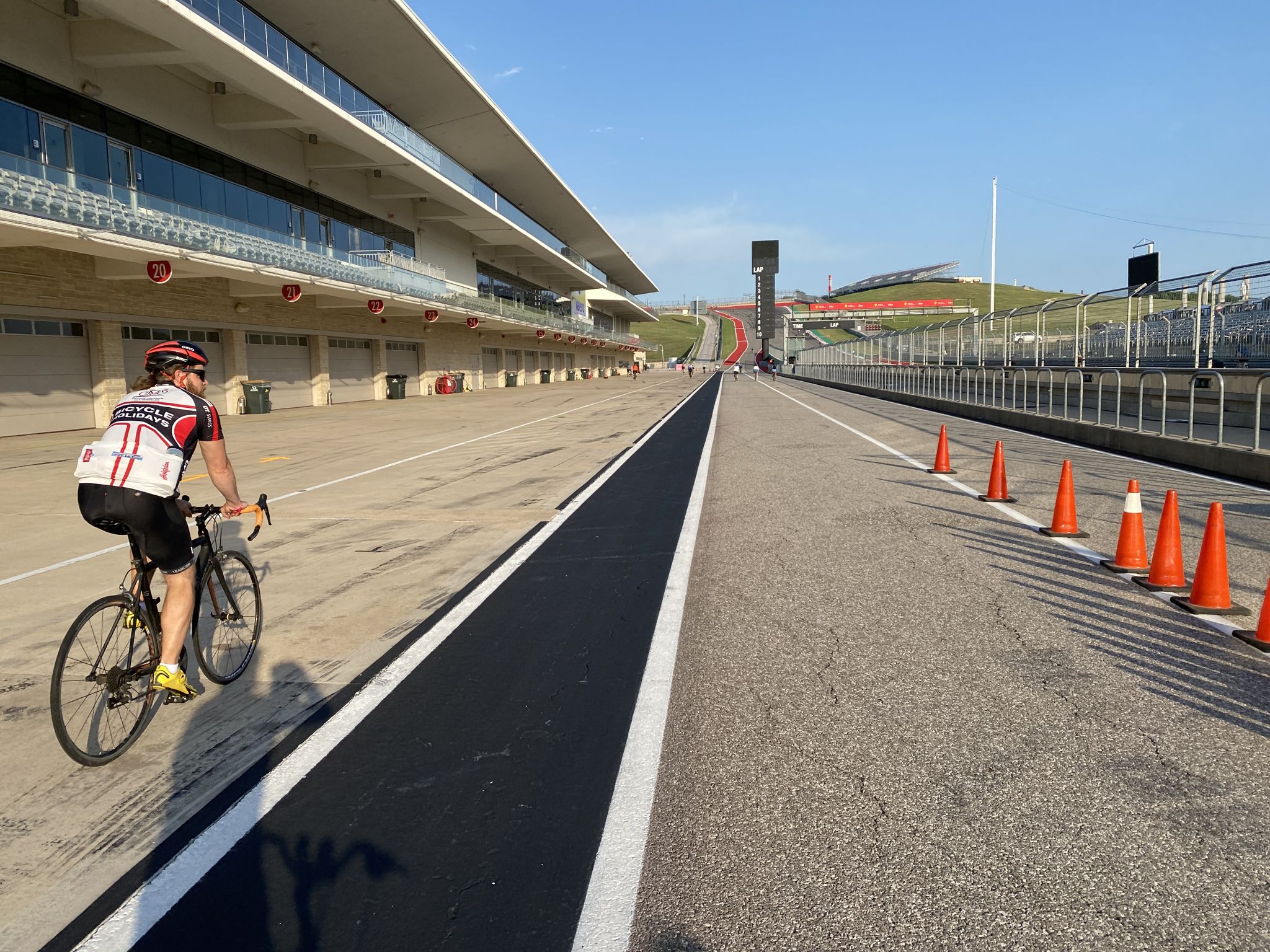 A person cycles on a wide paved track with buildings and orange cones. - COTA Track Loop
