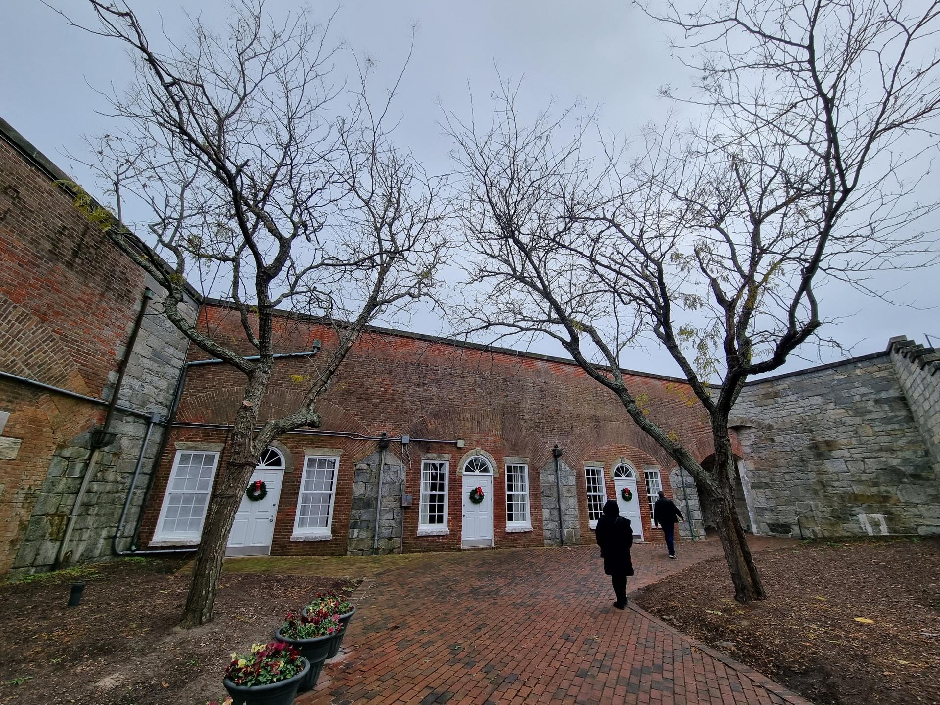 Two people walk past a brick building with white doors and bare trees.