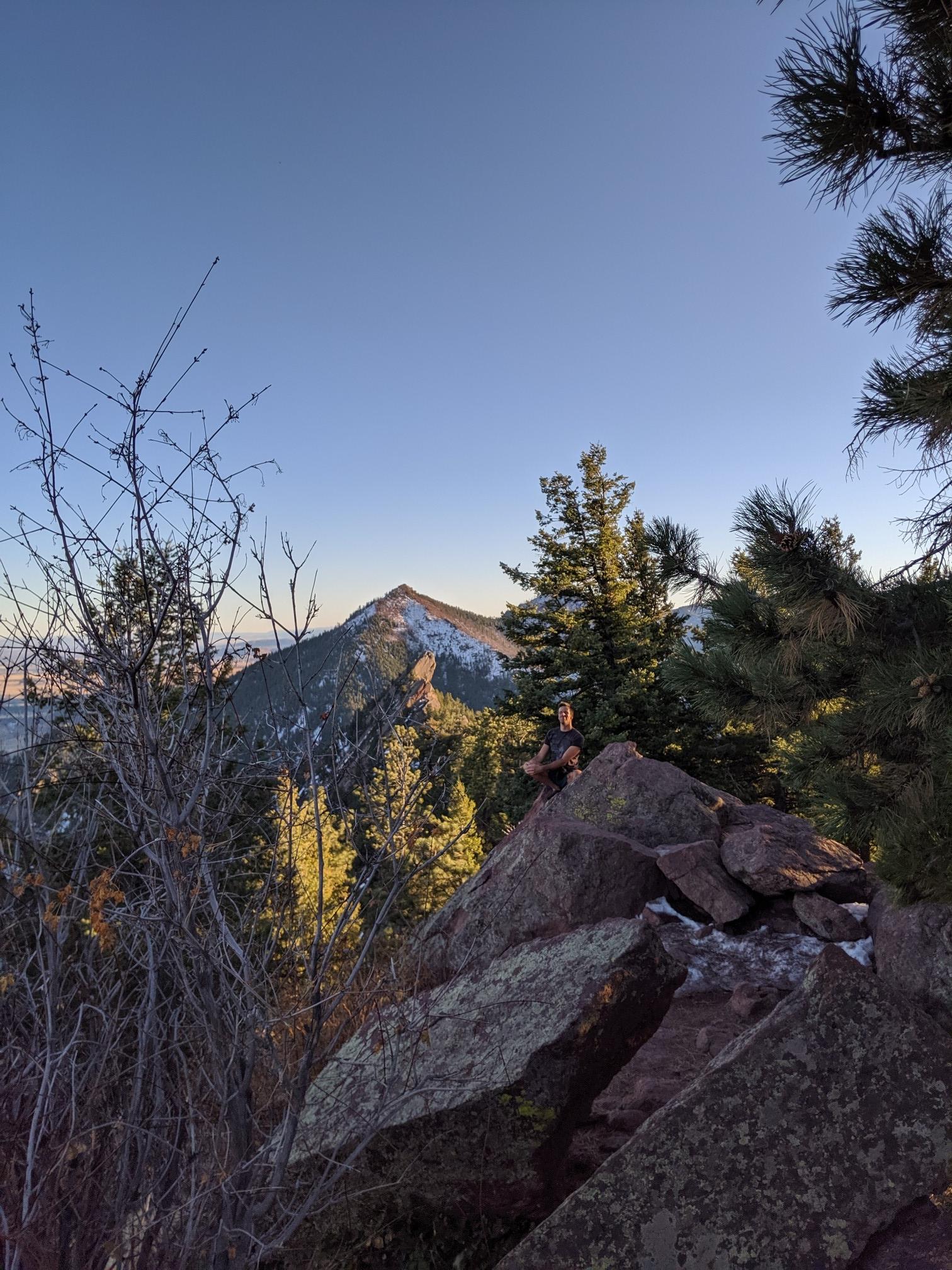 Mountain peak at sunrise, framed by pine trees and rocks, under a clear sky.