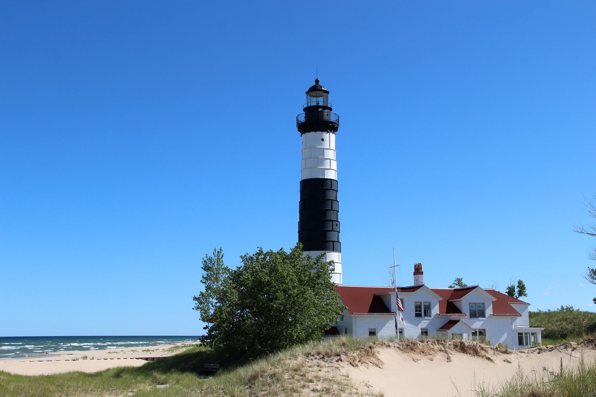 A tall black and white lighthouse next to a white building on a sandy beach. - Lighthouse Trail Loop