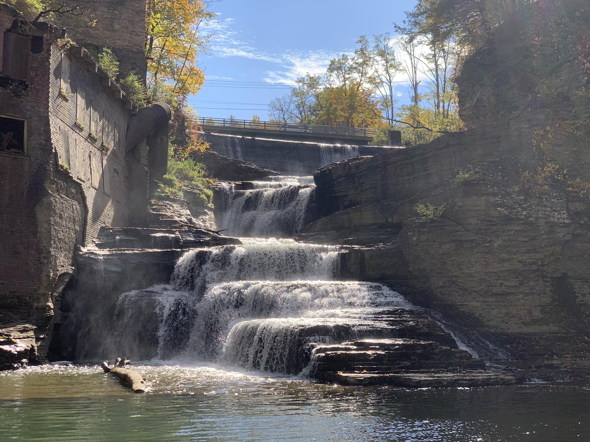Sunny view of a multi-tiered waterfall cascading past old structures and autumn trees.