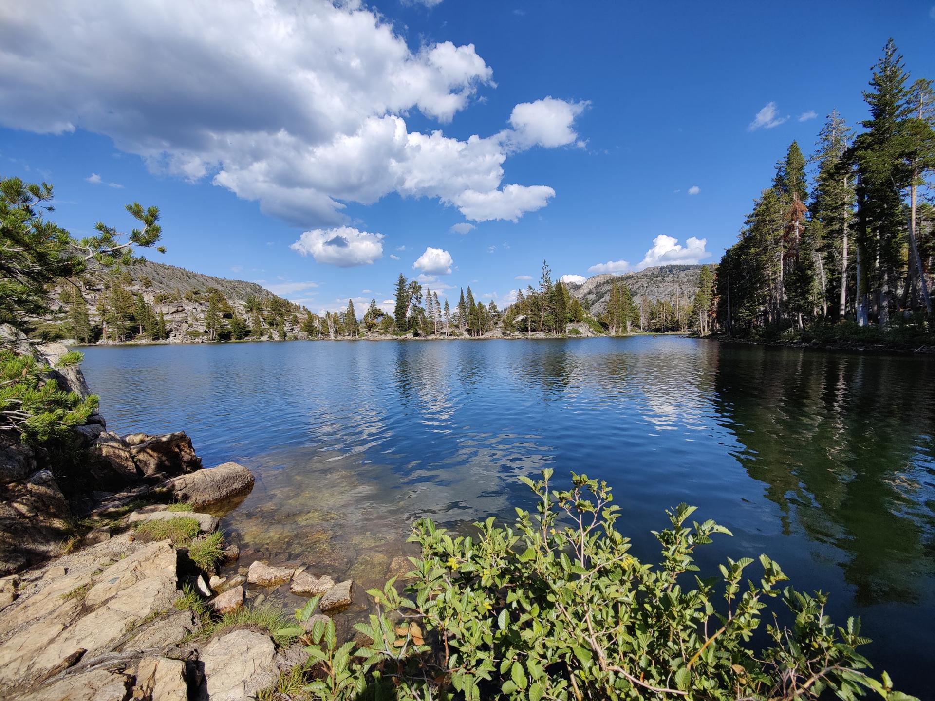 A wide, calm lake reflecting blue sky and white clouds, surrounded by forests.