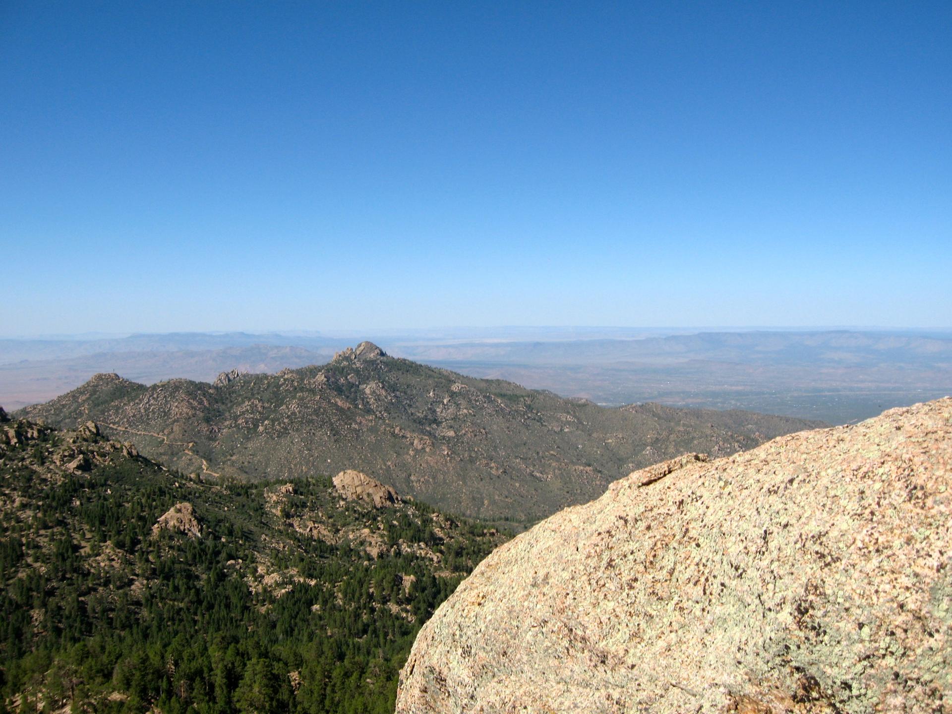 Sunny view of forested mountains and distant hazy peaks from a rocky foreground.