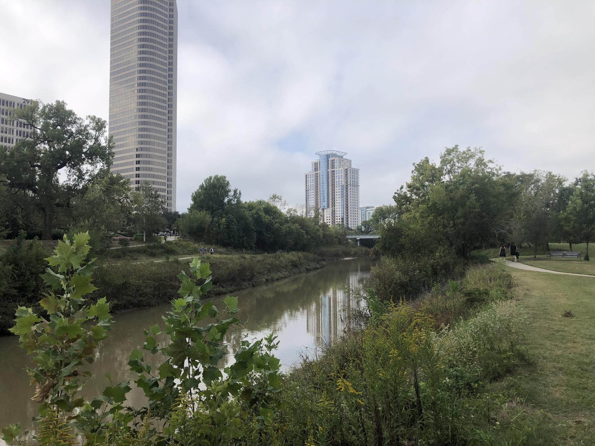 Calm river through green banks with tall city buildings under an overcast sky.