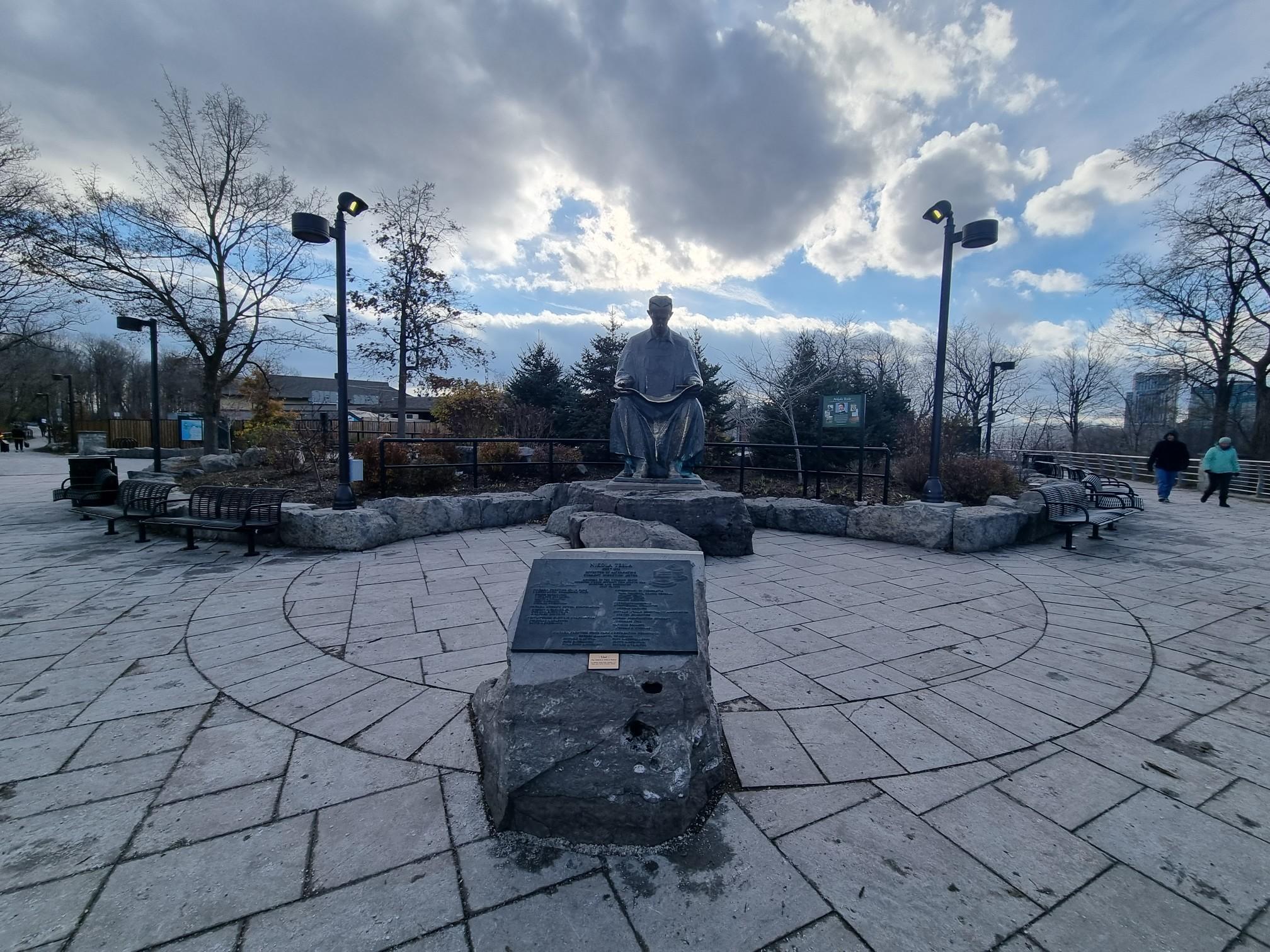Stone statue in a paved plaza with distant people under a cloudy sky.