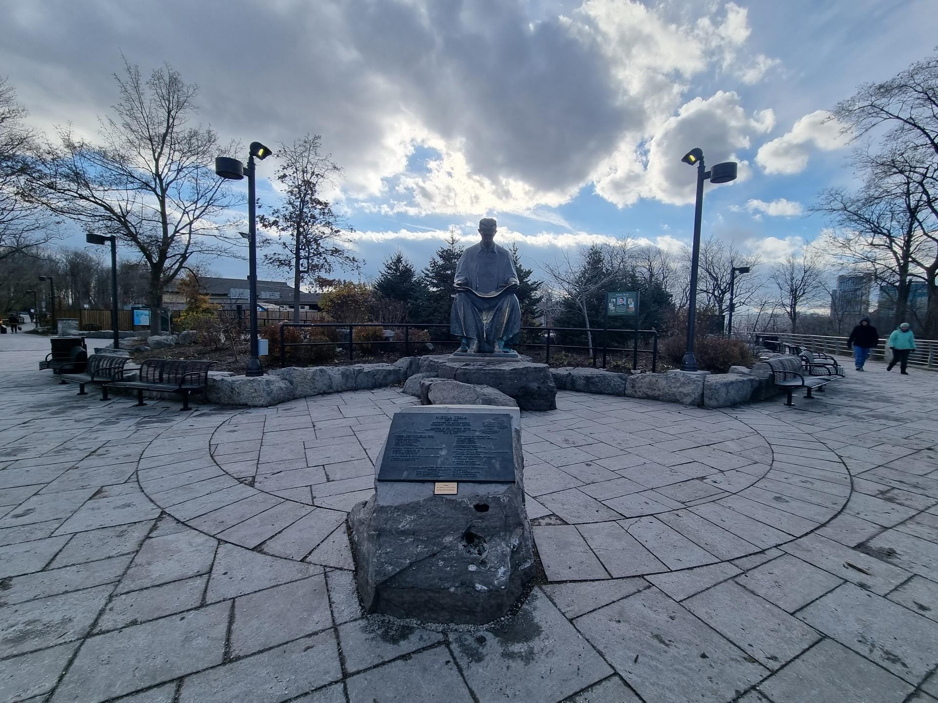 Stone statue in a paved plaza with distant people under a cloudy sky.