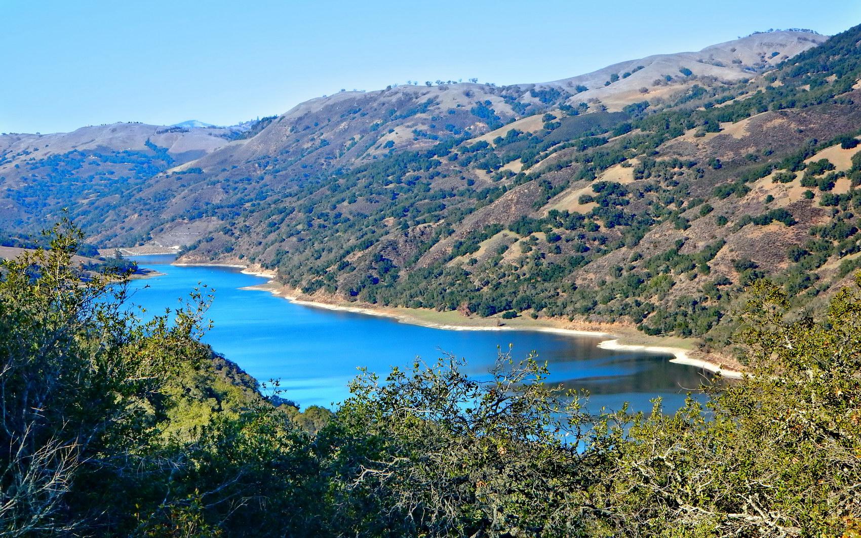 Bright blue lake surrounded by green and brown hills under clear sky.