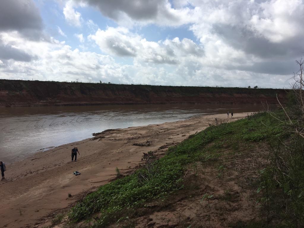 People walk along a wide sandy riverbank under a cloudy sky.