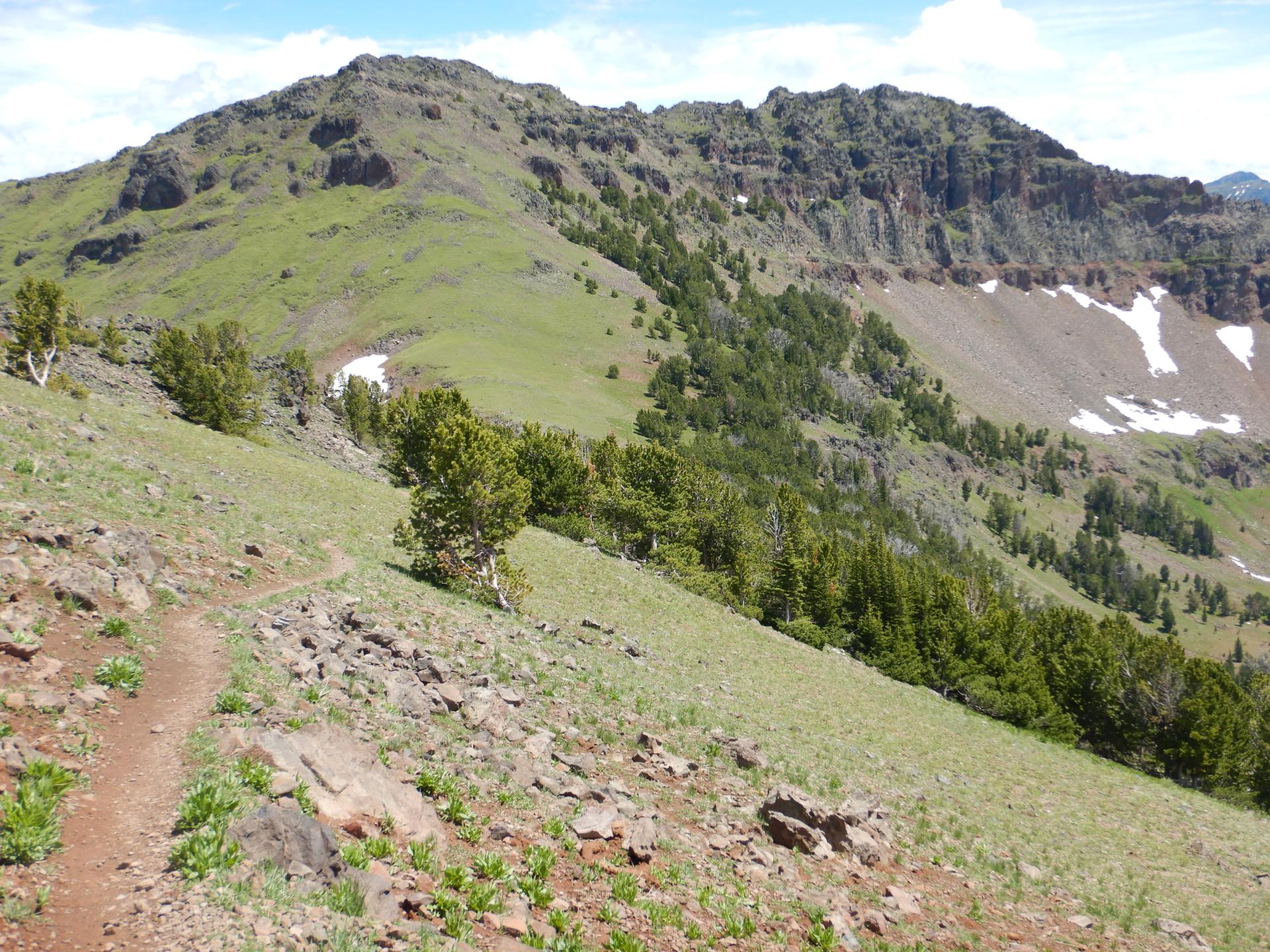 A dirt trail traverses a green mountain slope with rocky peaks and snow patches.