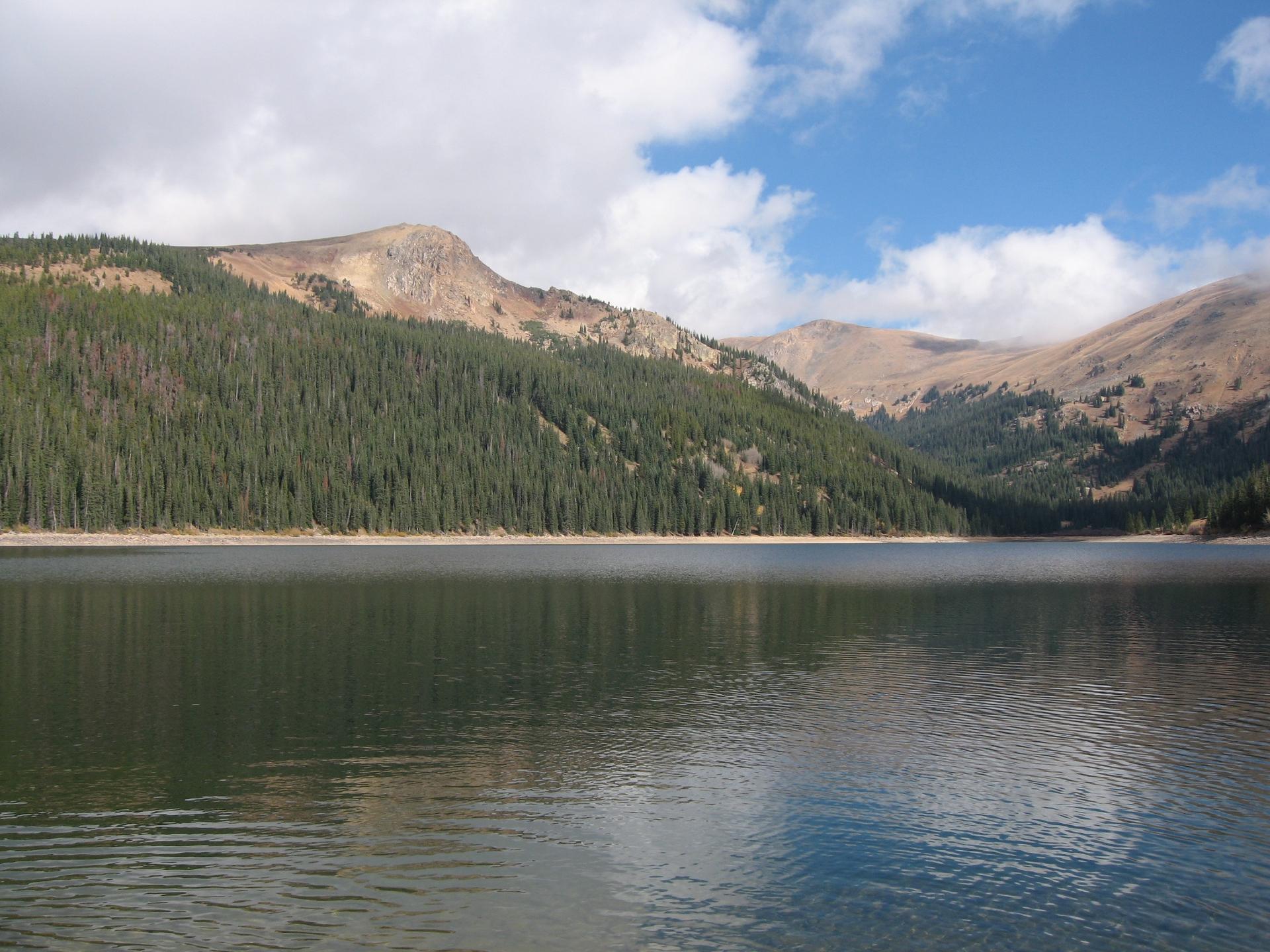 Vast lake reflecting sky, surrounded by forested and rocky mountains under a partly cloudy sky.