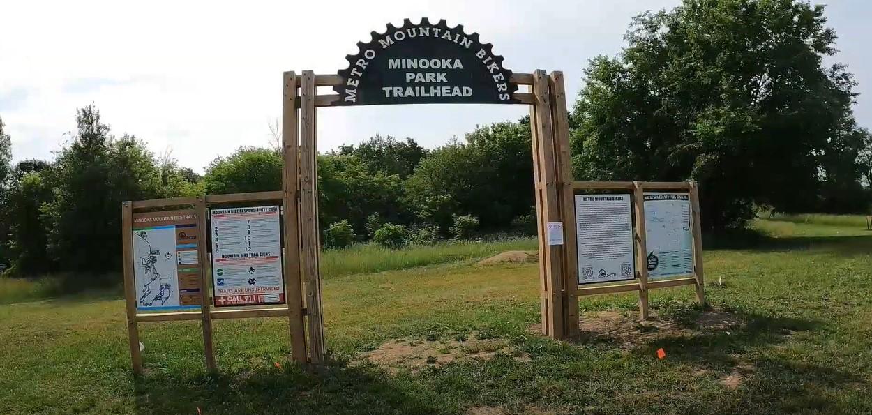 Wooden trailhead archway with information boards in a grassy field under daylight.
