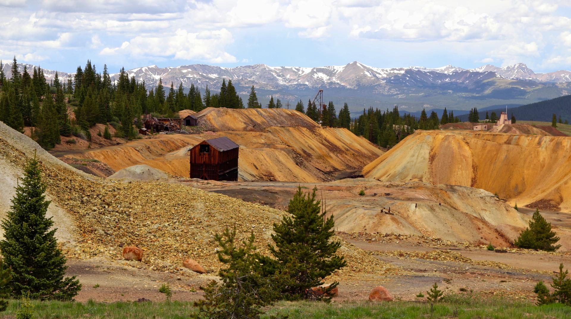 Mining landscape with orange tailings, old buildings, evergreen trees, and snowy mountains.