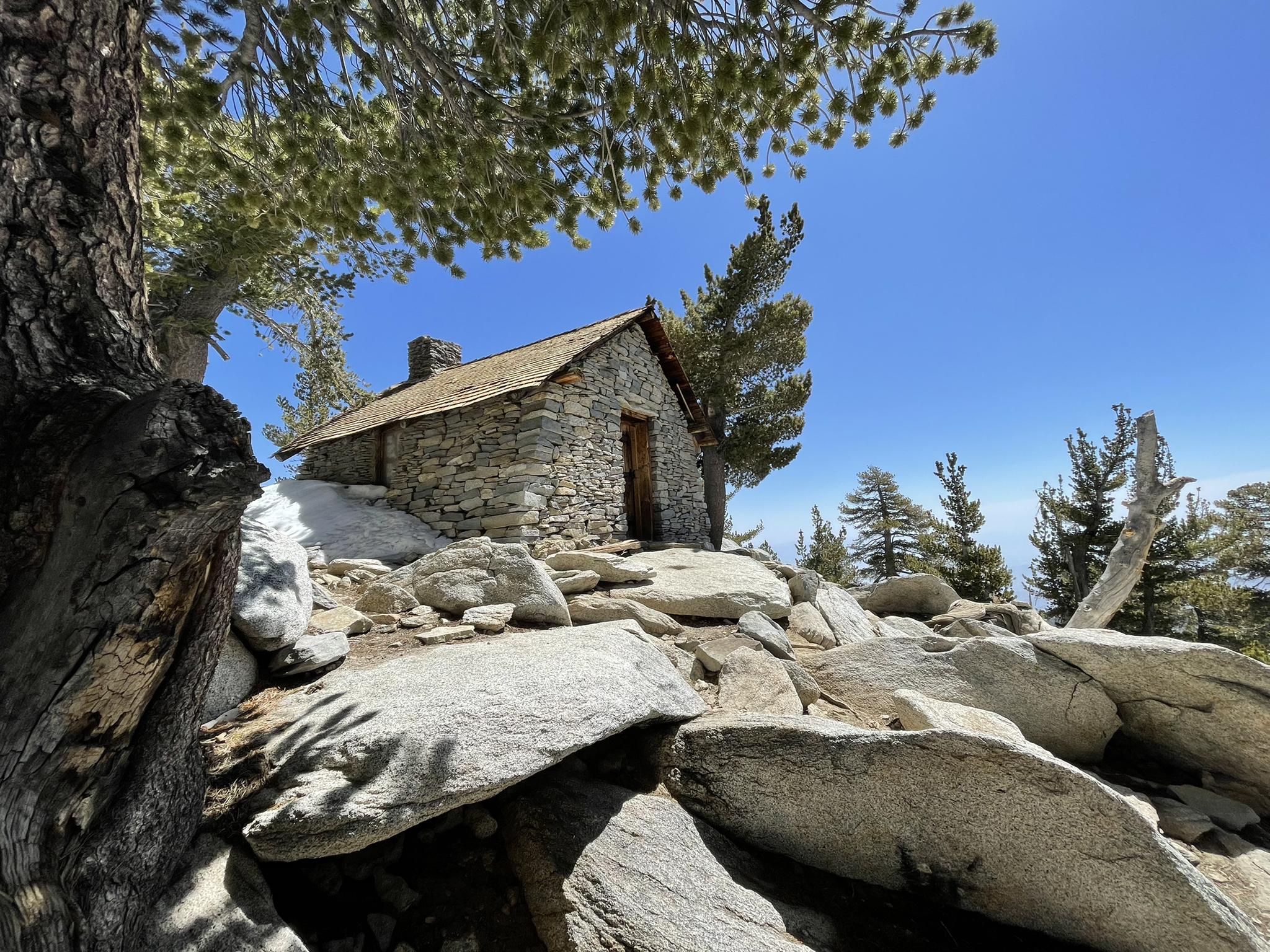 A rustic stone cabin sits atop a rocky, tree-covered peak under a clear sky.