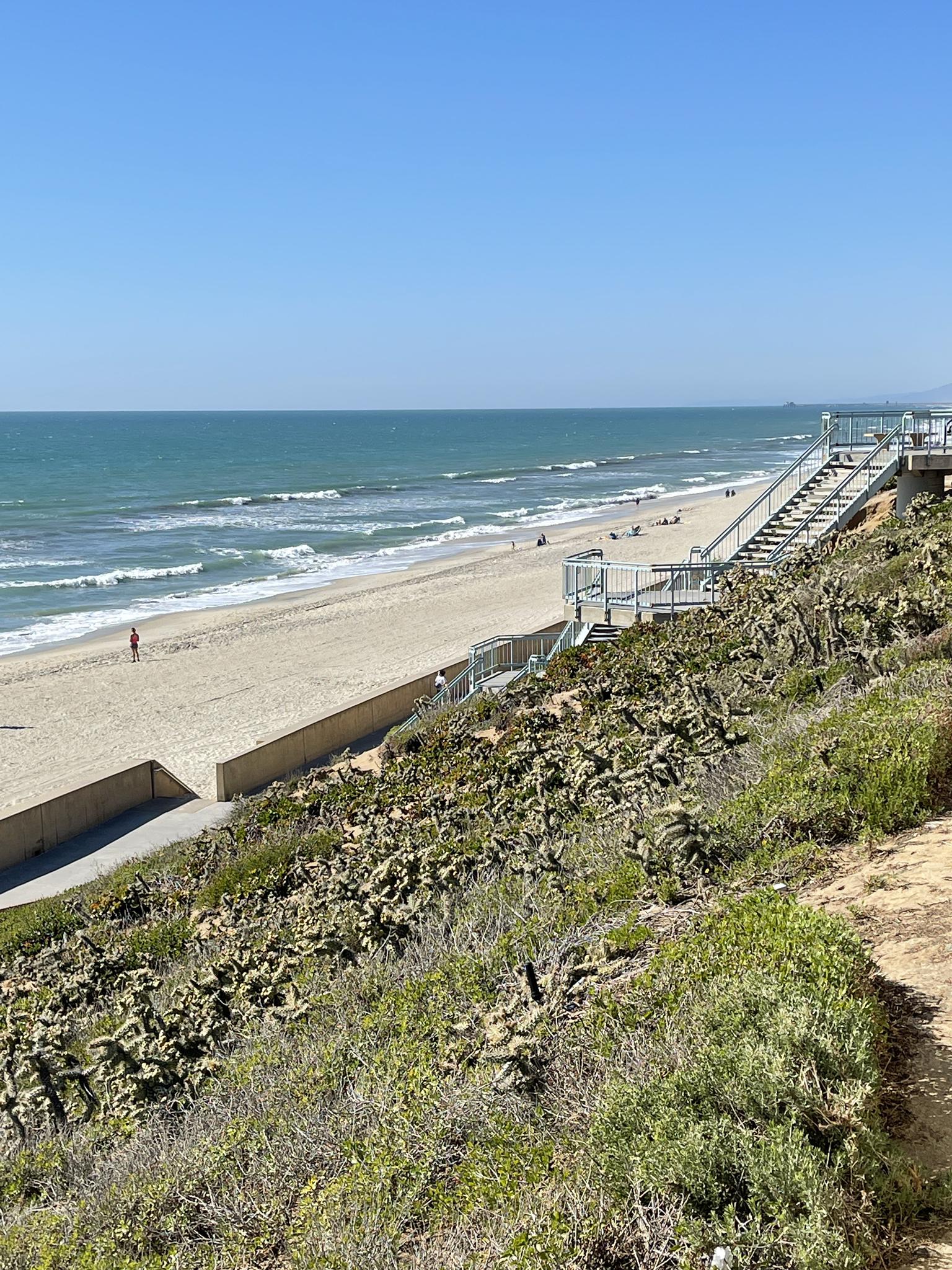 Sunny beach with ocean waves, sandy shore, and stairs descending from a green hillside.