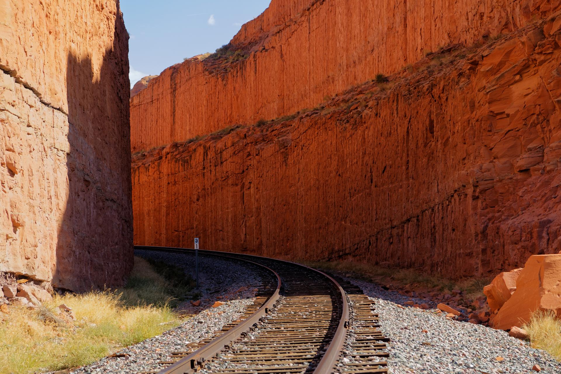 Train tracks curve through a deep, narrow red rock canyon under bright light.