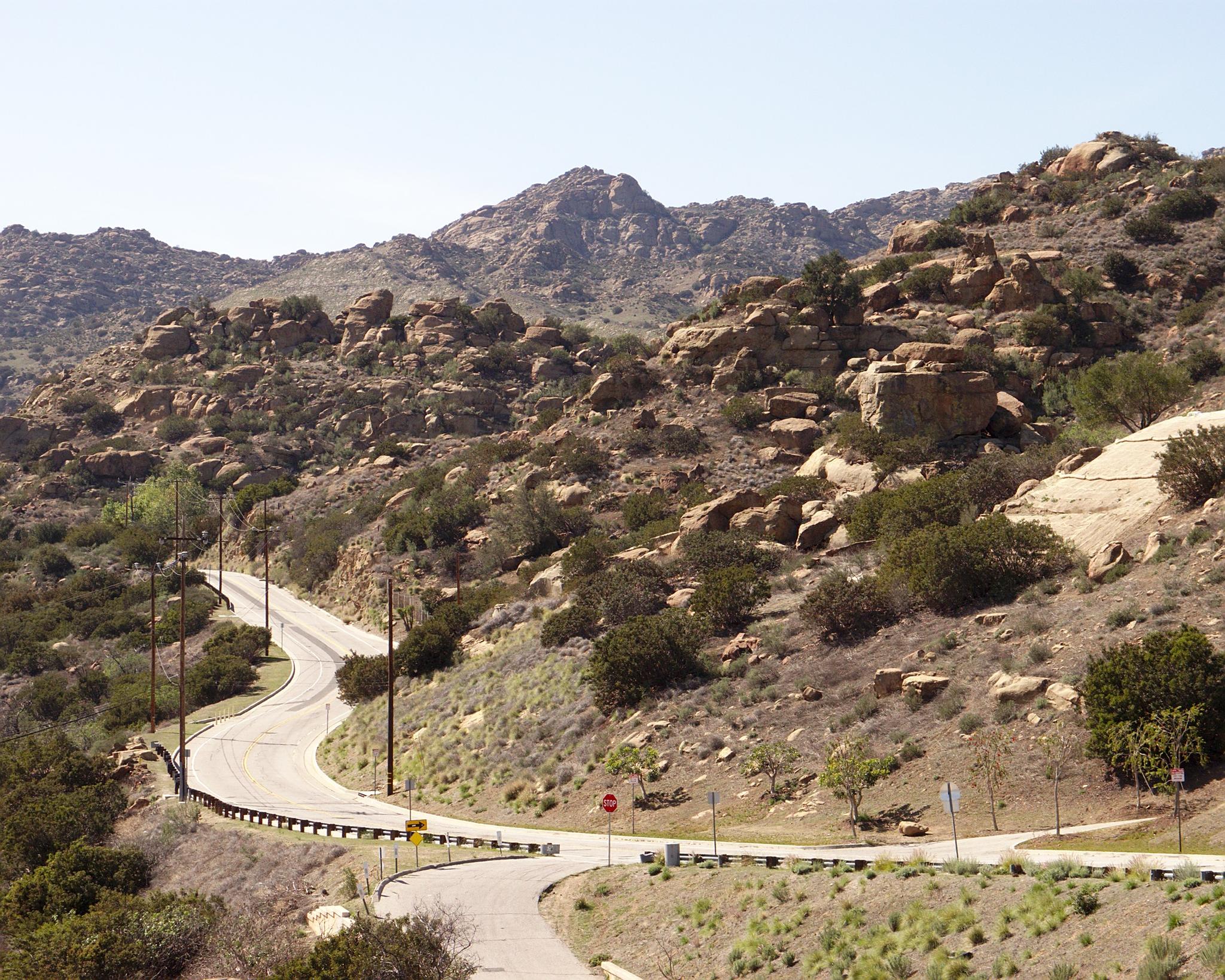 A winding road cuts through rocky, brush-covered mountains on a sunny day.