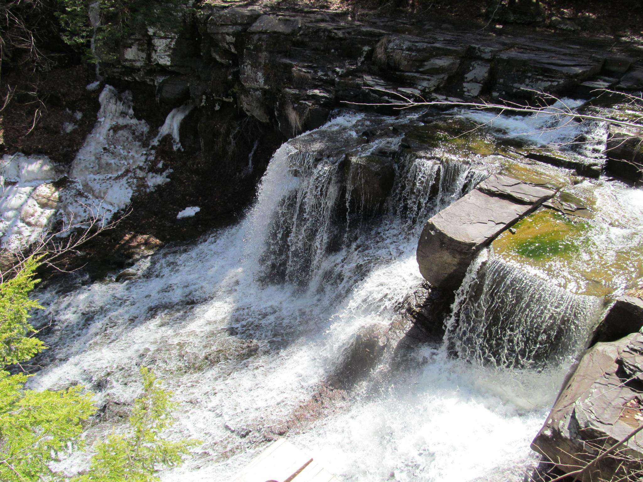 Powerful waterfall cascades over dark rocks with some white snow patches.