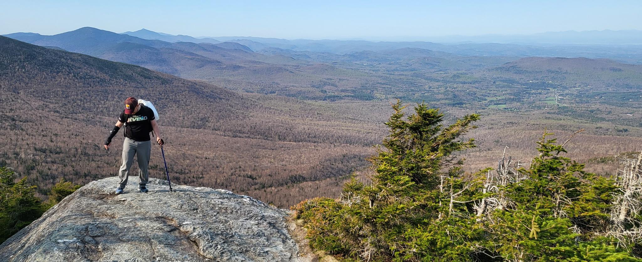 Hiker with backpack and poles stands on a rocky peak overlooking a vast valley.