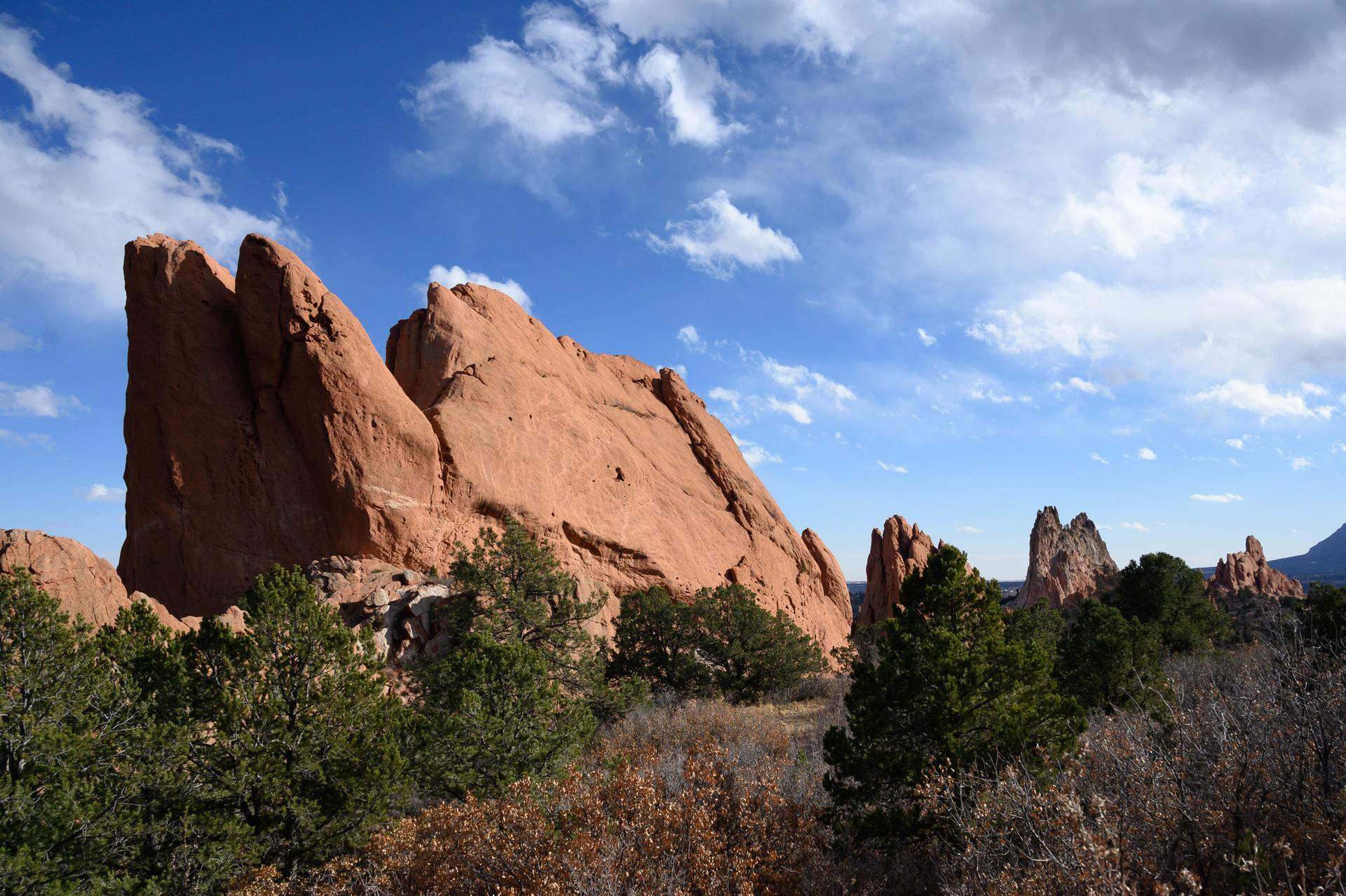 Large red rock formation dominates the left, with smaller rocks and trees under a blue, cloudy sky.