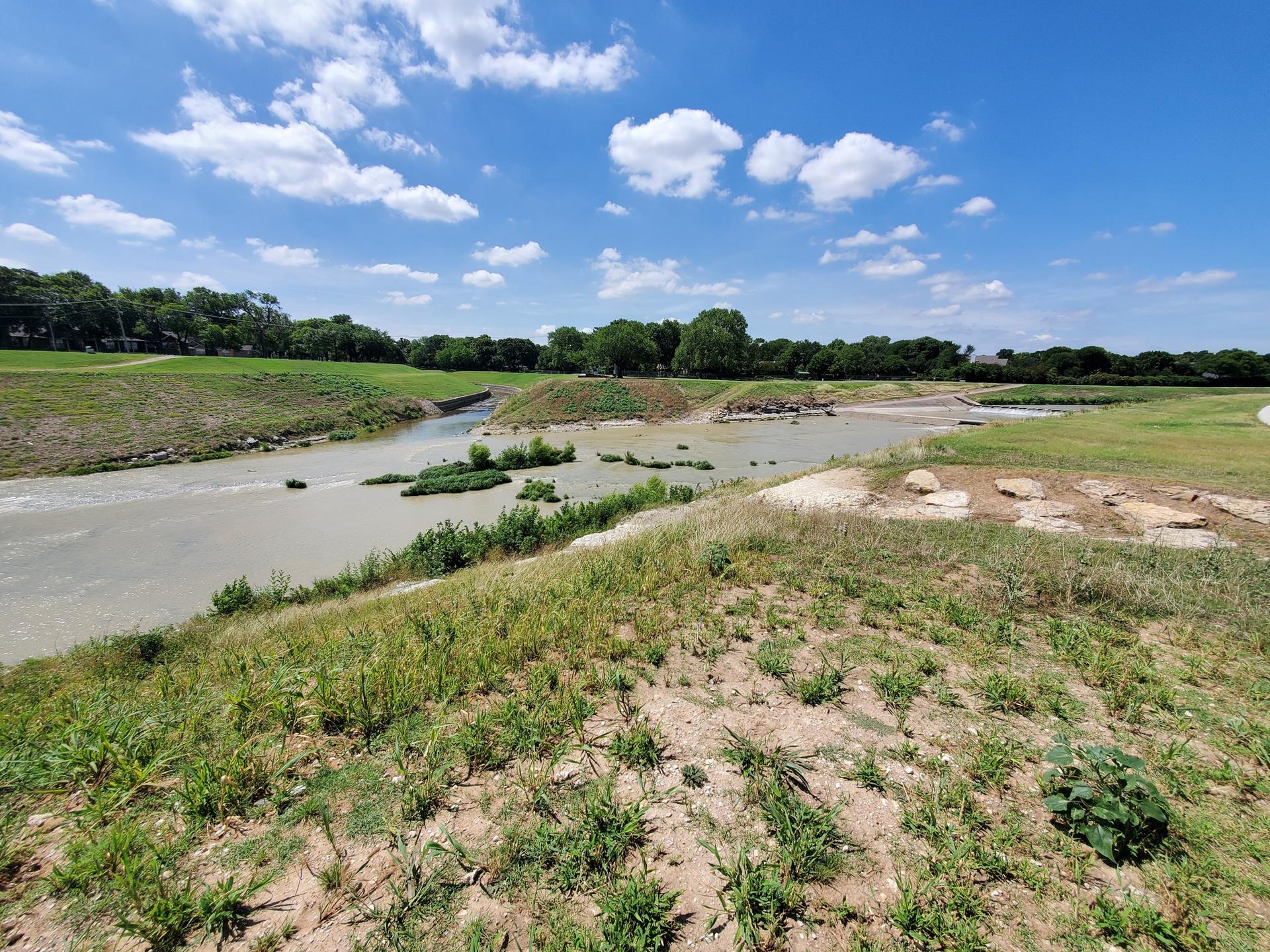 Wide muddy river flowing through grassy banks under a bright blue sky.