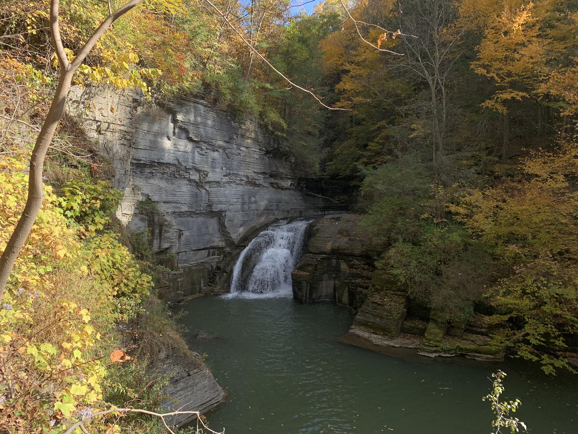 Sunny view of a waterfall plunging into a pool, surrounded by autumn trees.