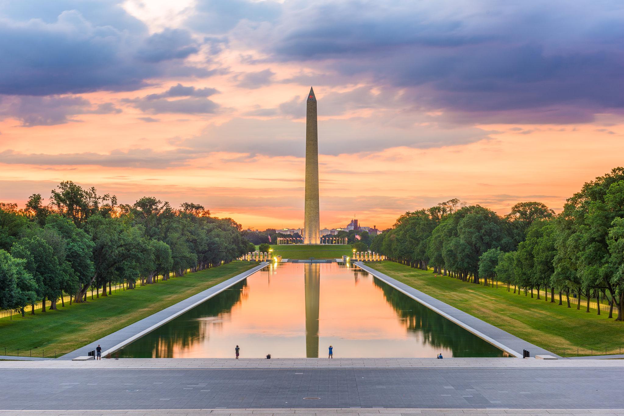 Obelisk reflecting in long pool at sunset, green trees, people visible.