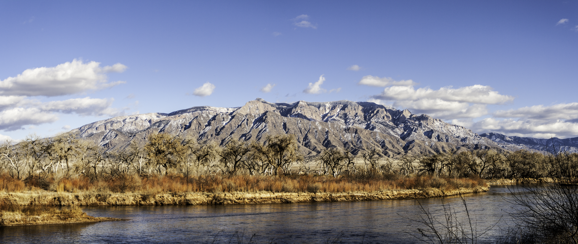 Panoramic view of a wide river, bare trees, and a snow-dusted mountain range.