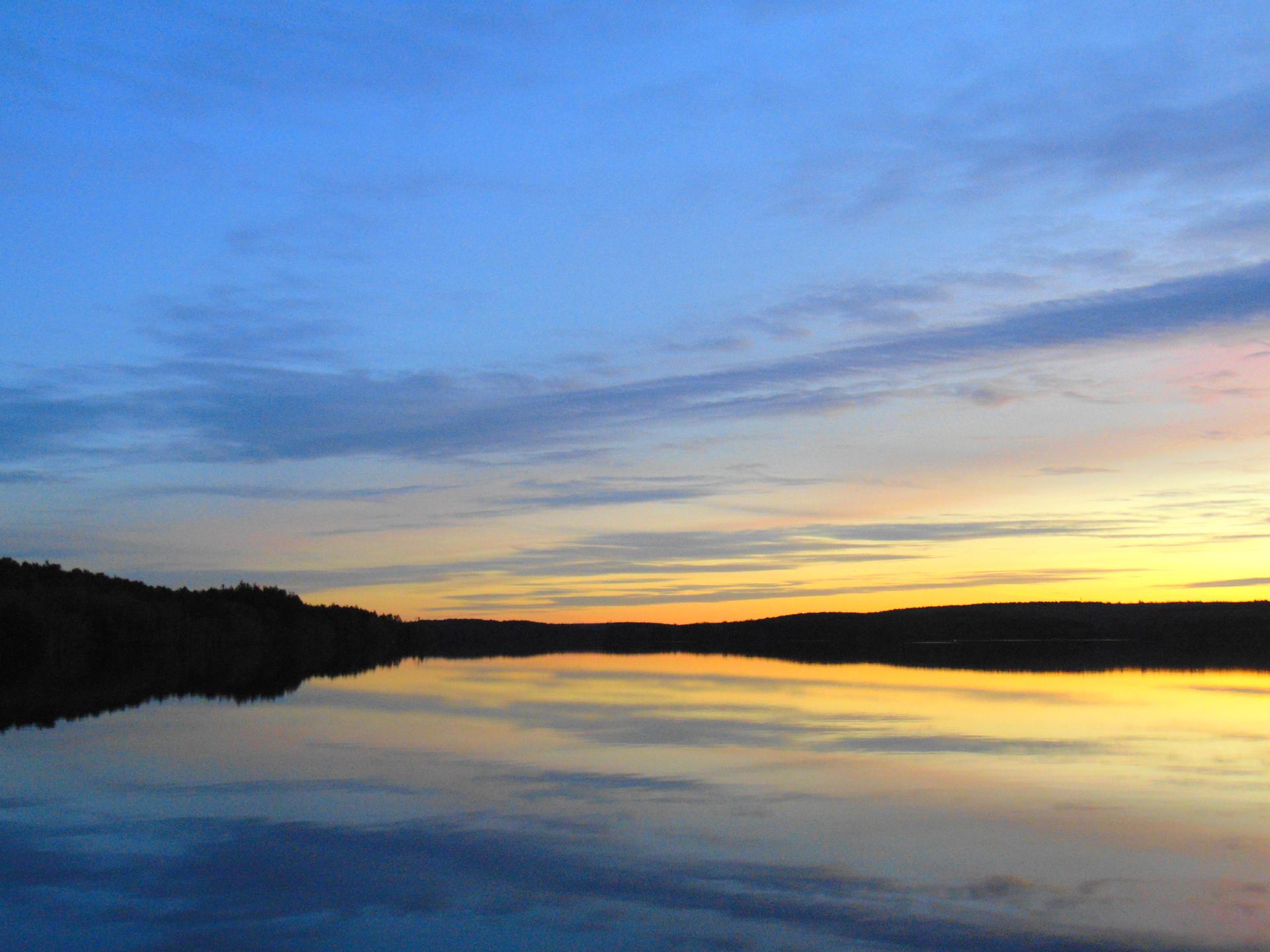 Vibrant sunset sky reflecting on calm water with a dark forested horizon.