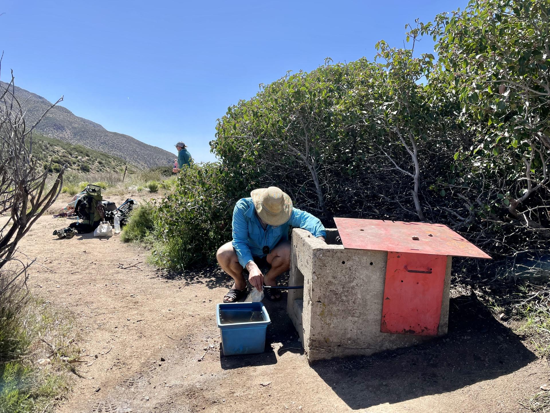 Person crouching by concrete water box, another person in sunny desert background.
